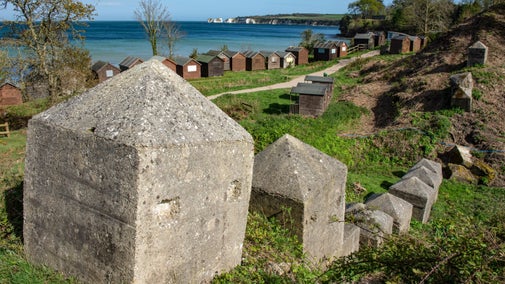 Concrete blocks, part of World War II defences, poke out of the grass, with a row of beach huts in the background at Middle Beach, Studland Bay, Dorset. Across the blue sea in the bay are the white cliffs of Old Harry Rocks